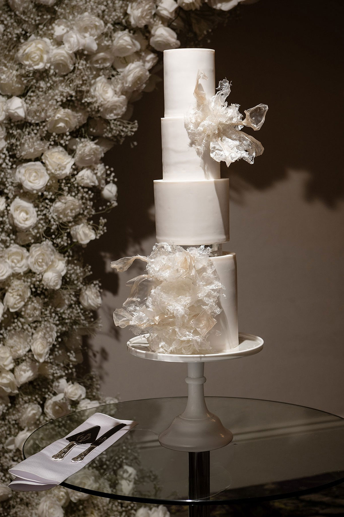 Modern white tiered wedding cake with sculptural sugar details beside a white rose flower wall at Old Course Hotel wedding in St Andrews.