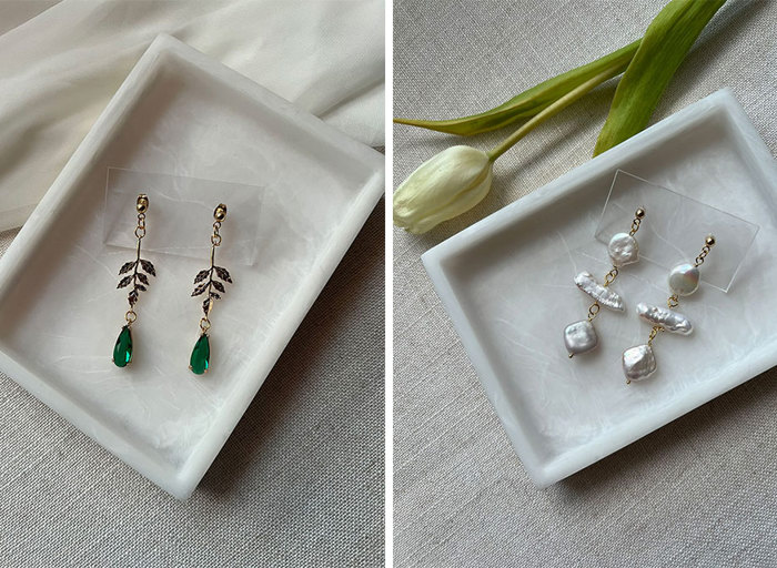 a pair of green stone and leaf detail drop earrings on a marble tray on left. A pair of abstract-shaped pearl drop earrings on a marble tray