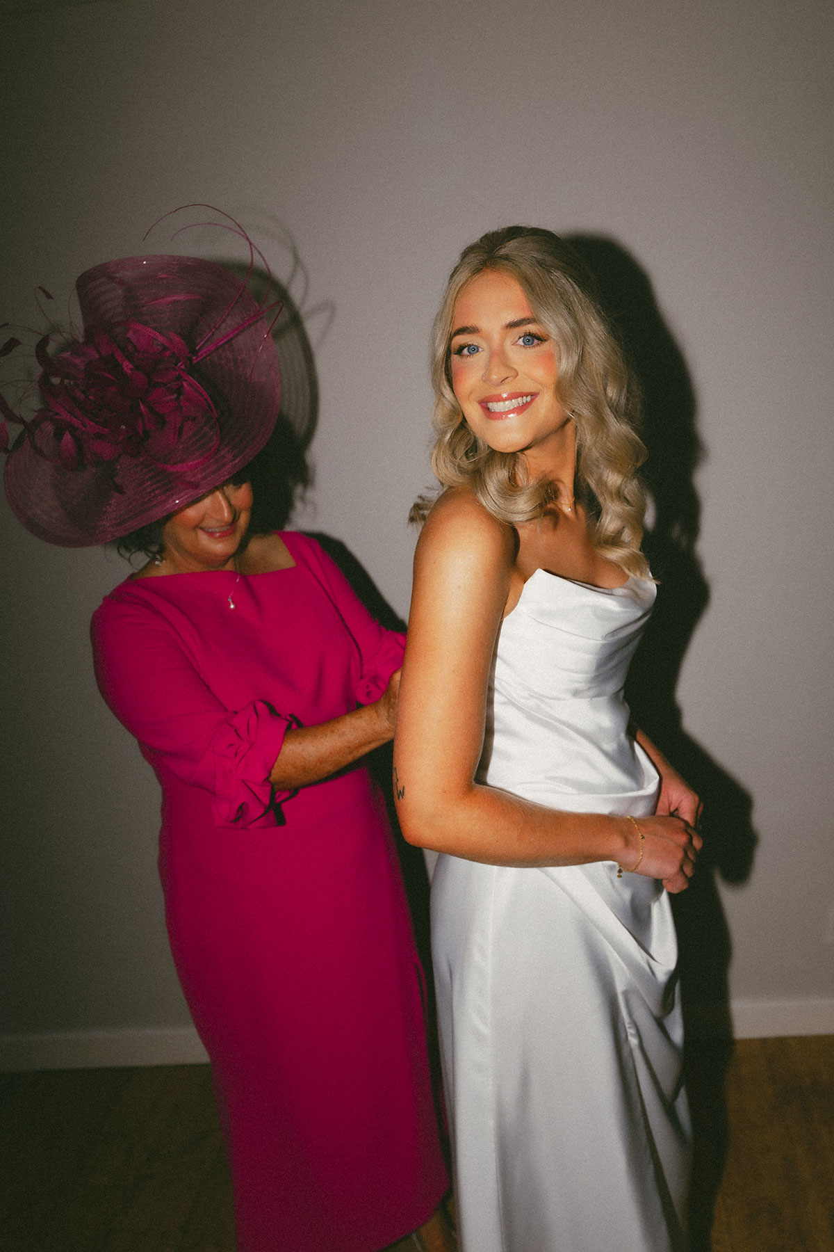 Older woman in a bright pink dress and large fascinator fastens the back of a bride’s white satin gown as the bride looks over her shoulder and smiles