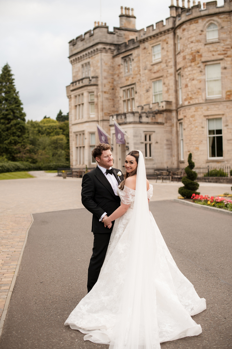 Bride and groom posing outside Crossbasket Castle, with the bride's long lace wedding dress and veil on display.