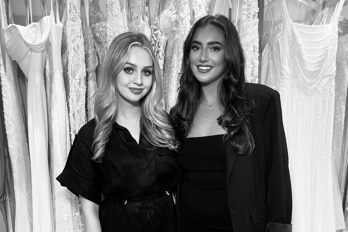 black and white image of blonde and brunette women stood next to each other, both wearing black tailored clothing, in front of a rail of wedding dresses