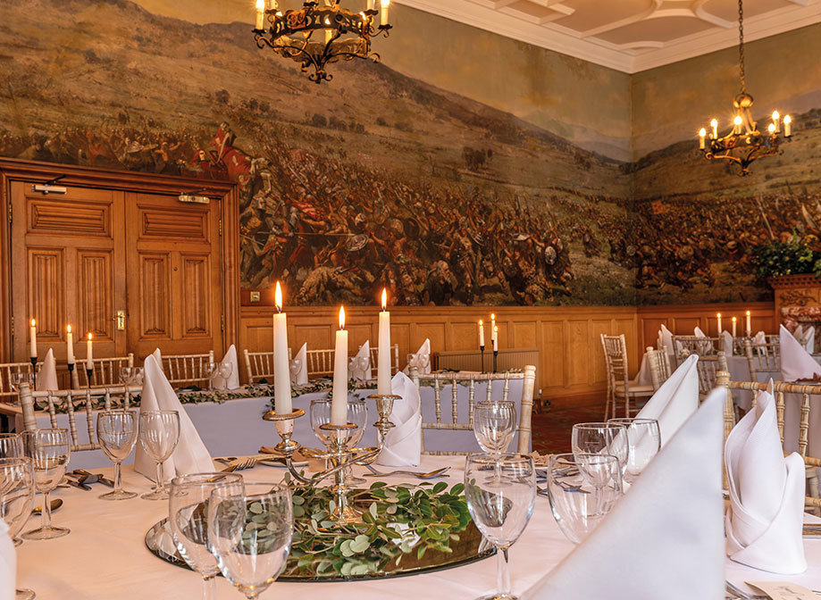 A room with a mural of the Battle of Bannockburn on the walls, with white round tables and lots of candle sticks