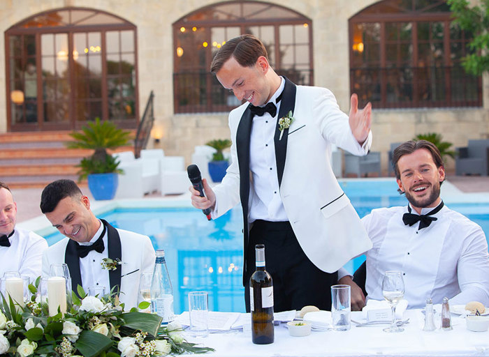 a smiling man holding a microphone. He is standing at a long table in front of a swimming pool as other men wearing bow ties sit either side