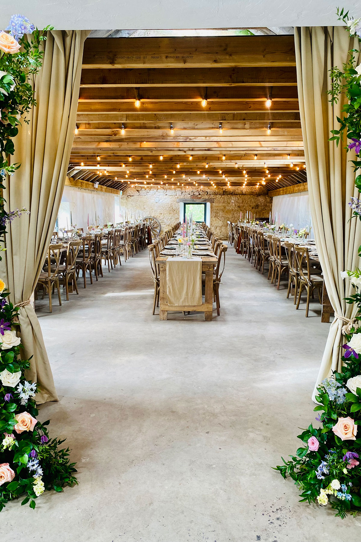 The Byre at Blackshaw Barns set for a wedding meal, with long wooden banquet tables, rustic chairs and string lights hanging from the exposed beams.