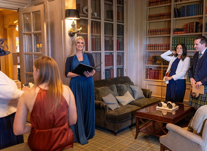 a woman wearing a long blue dress holding a black folio standing in a cosy looking room with bookcases and sofas as other people look towards her