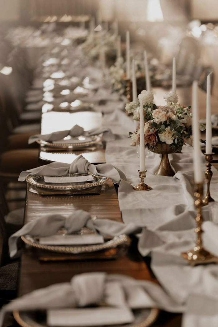 a long table set with white napkins and bronze plates and bronze candlestick holders