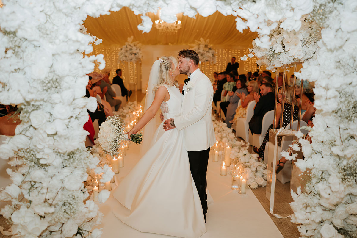 Newlyweds kissing at top of aisle framed by white floral arch and candles at Ingliston Country Club
