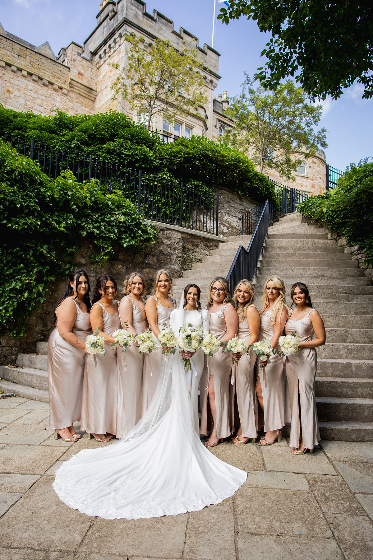 Bride in a long-sleeved white gown with a flowing train stands with her bridesmaids in champagne satin dresses, holding white bouquets on stone steps outside a Scottish castle