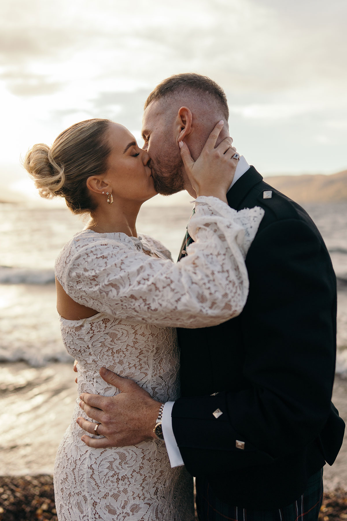 Bride in lace gown and groom in kilt share a kiss by the Scottish coast at sunset