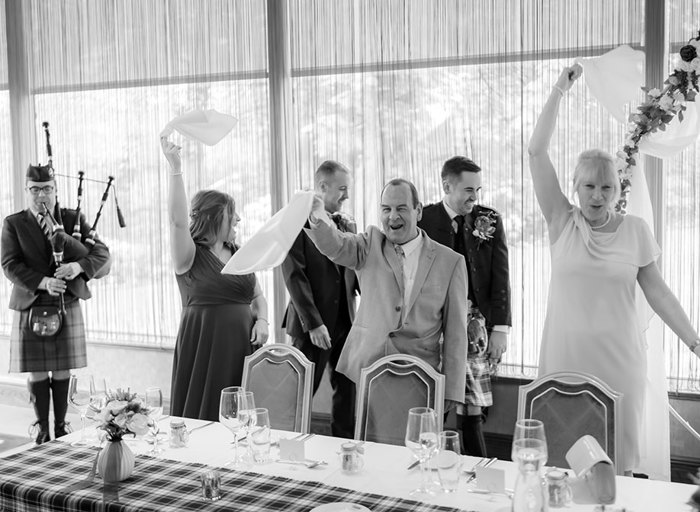 standing wedding guests swinging napkins as two grooms enter top table at a wedding. A bagpiper plays on left of image