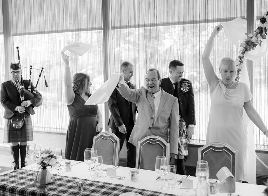 standing wedding guests swinging napkins as two grooms enter top table at a wedding. A bagpiper plays on left of image