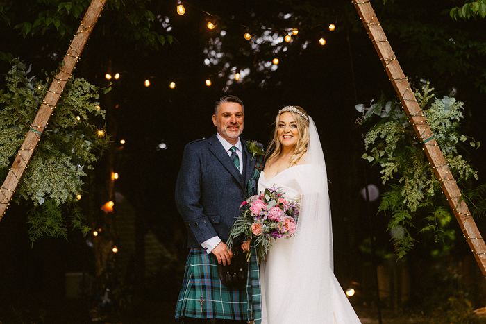 Couple standing in front of archway, bride holding flowers and groom wearing a kilt