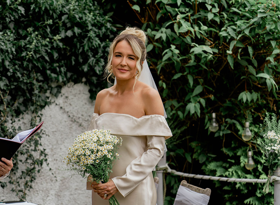 a bride wearing an off-shoulder long-sleeve ivory wedding dress holding a large bunch of daisies with greenery and white pebble-dash wall in background