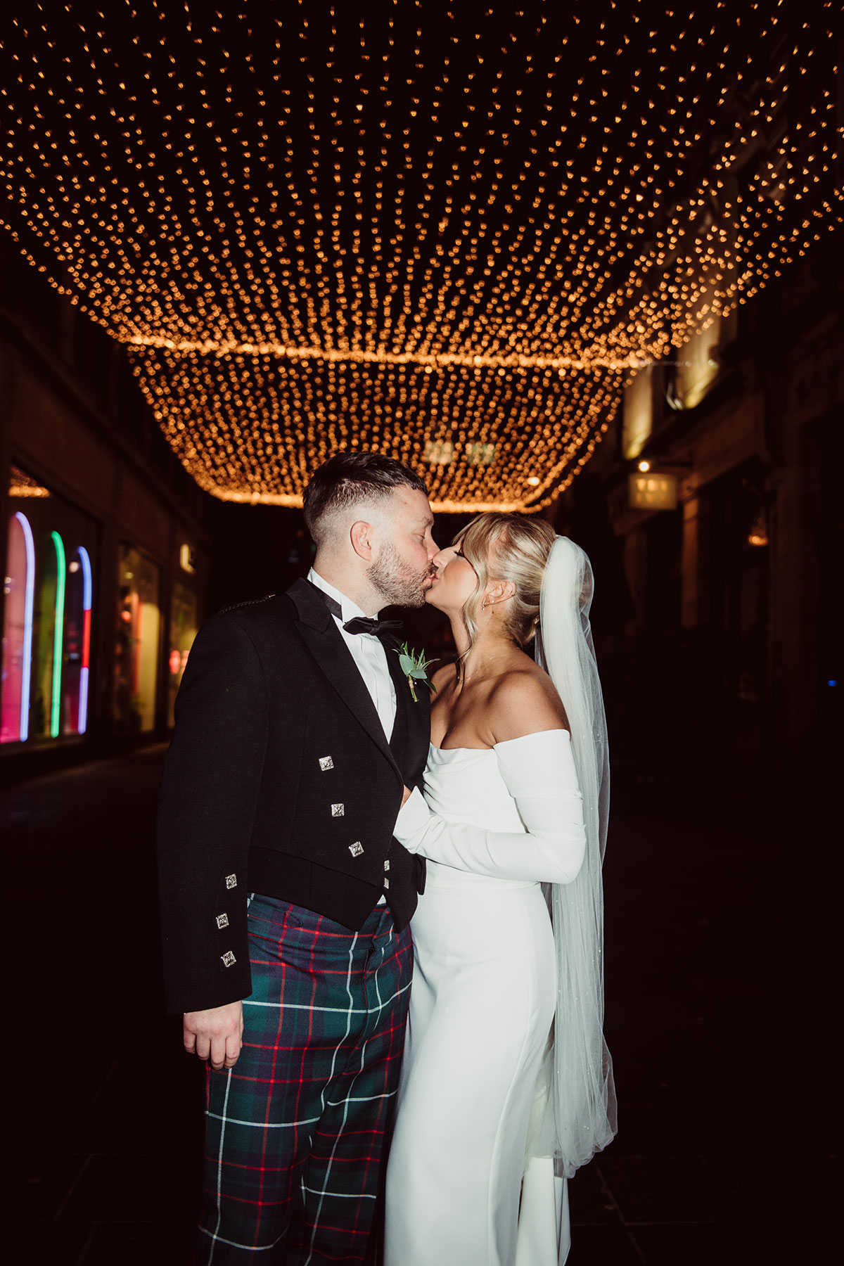 Bride and groom kissing outdoors at night beneath a canopy of warm white string lights