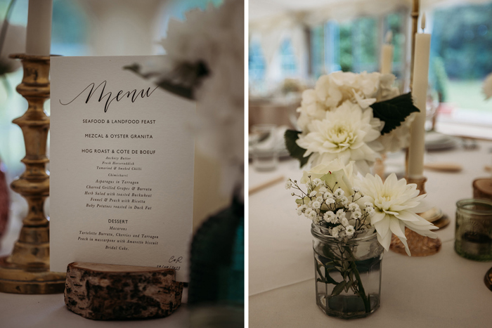 A Wedding Menu Propped On A Cut Log On Left And White Flowers In A Jam Jar On A Table On Left