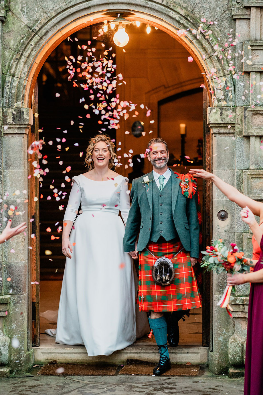 bride in white wedding dress and veil and groom in red and green tartan kilt step out of arched building opening hand in hand as pink heart shaped confetti is thrown on them