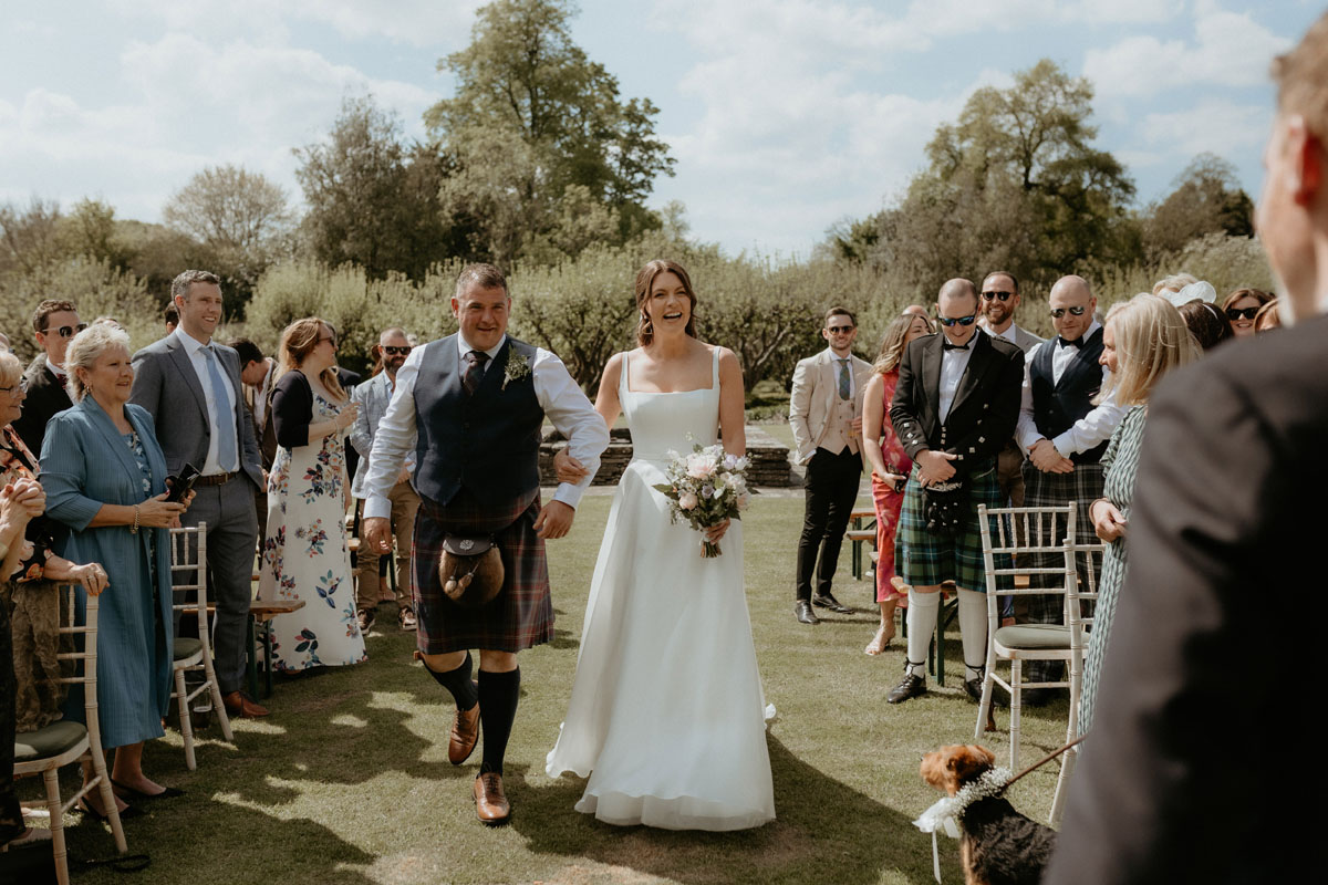 Bride walking down the aisle with a man in tartan at an outdoor wedding ceremony at The Byre at Inchyra.