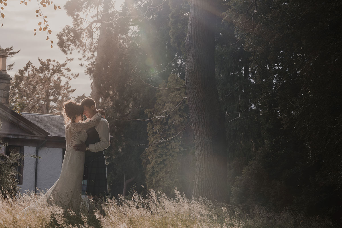 bride in long sleeve wedding dress stands face to face with groom in full kilt outfit with her arms around his neck and stood in front of white cottage and next to tree-filled woods in dimmed light