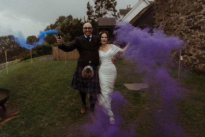 groom in blue, grey and white tartan kilt outfit holds out a blue smoke stick to one side while bride to his other side, whom he has an arm around, holds out a purple smoke stick to her other side