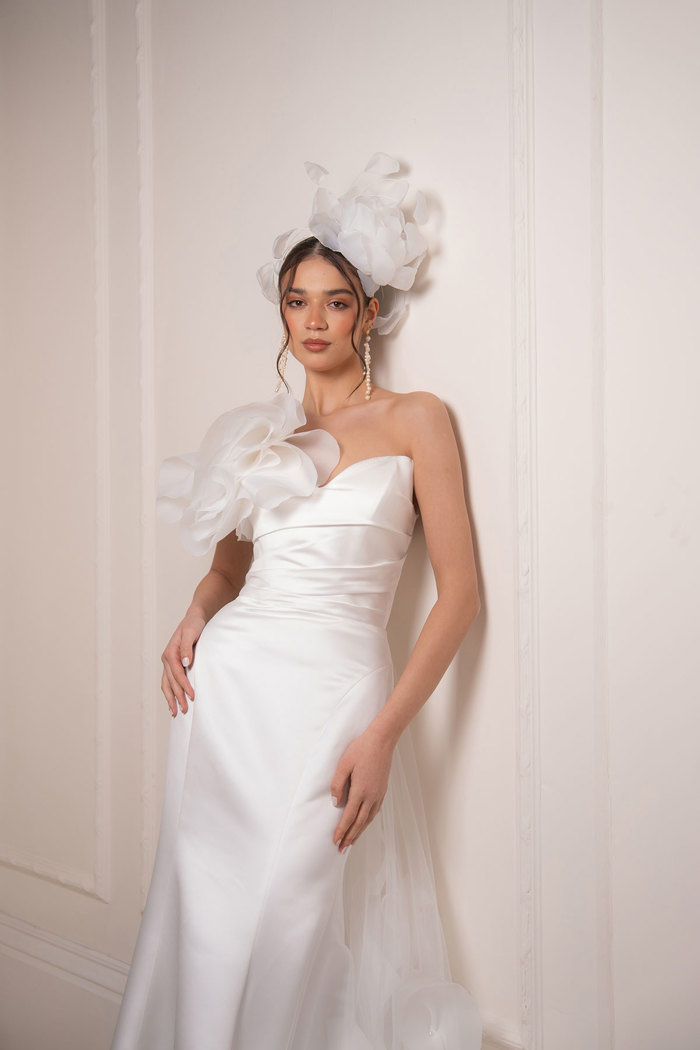 bride with dark brown hair wears a strapless ballgown wedding dress with large flower detail on one upper side of the bodice that matches the large floral headpiece as she stands in front of a white wall
