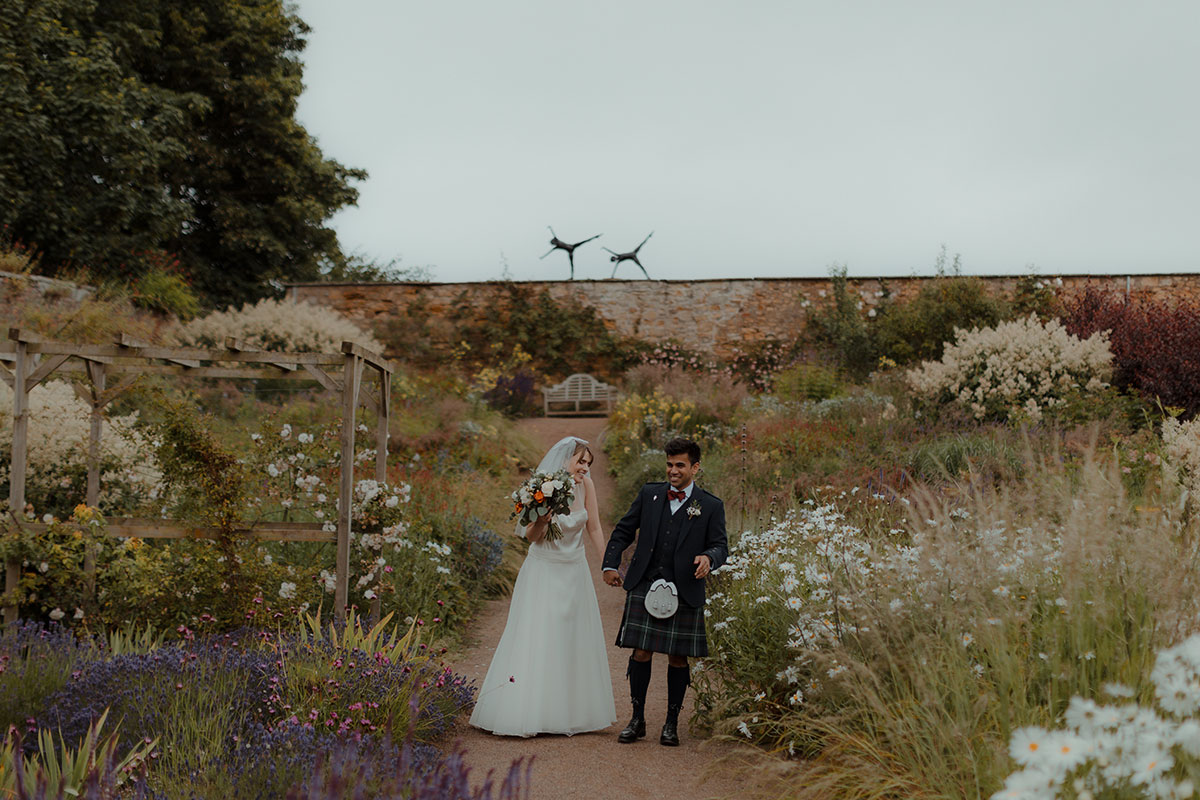 Bride and groom holding hands while walking through Cambo Estate’s flower-filled gardens, with pergola and stone wall in the background