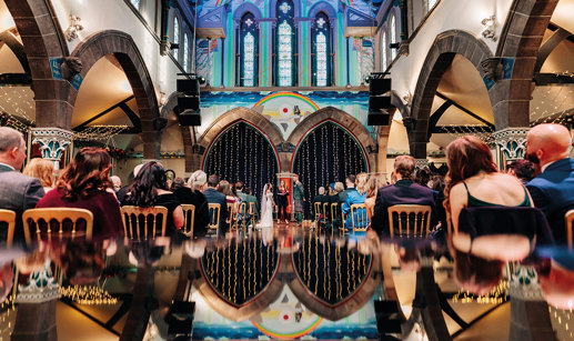 bride, groom and celebrant are seen at the top of a reflective aisle inside a large colourful arched space as rows of guests sit facing ahead, watching the couple
