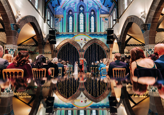 bride, groom and celebrant are seen at the top of a reflective aisle inside a large colourful arched space as rows of guests sit facing ahead, watching the couple