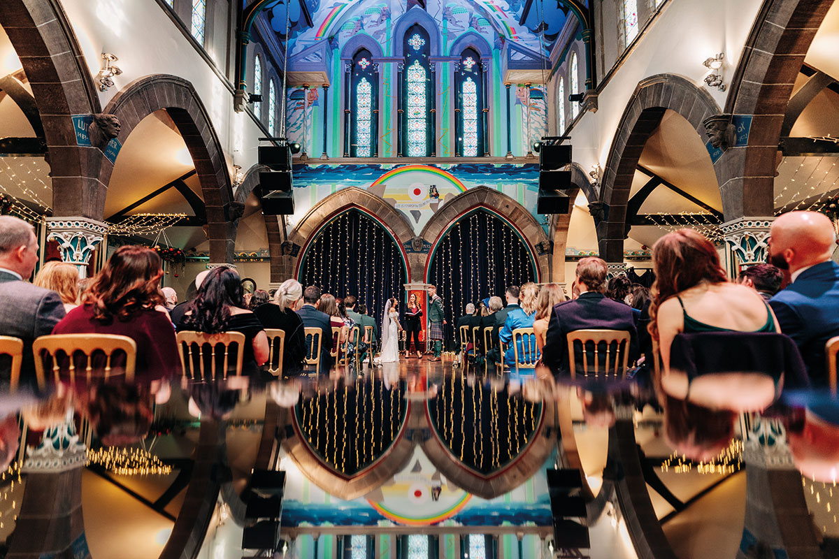 bride, groom and celebrant are seen at the top of a reflective aisle inside a large colourful arched space as rows of guests sit facing ahead, watching the couple