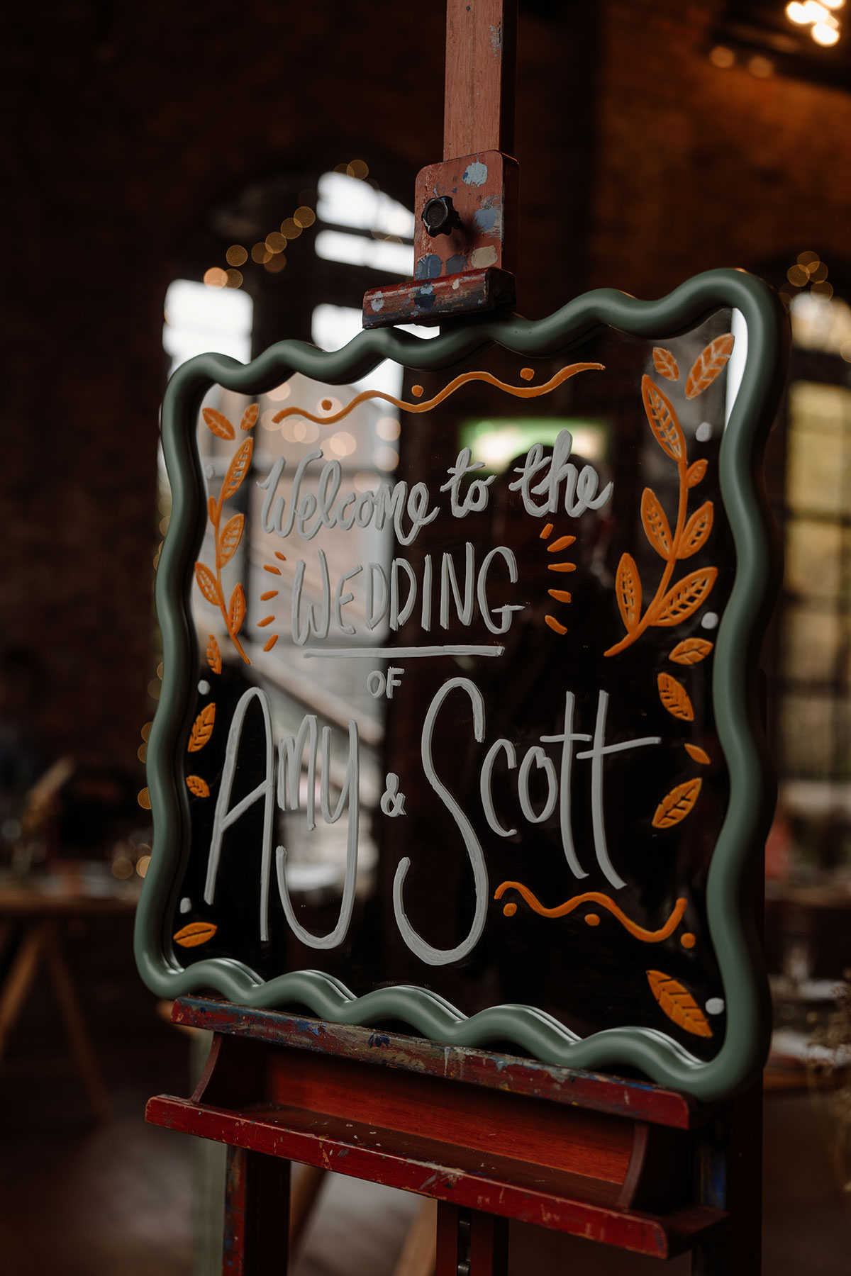Hand-painted wedding welcome sign displayed on an easel inside an industrial wedding venue decorated for a reception