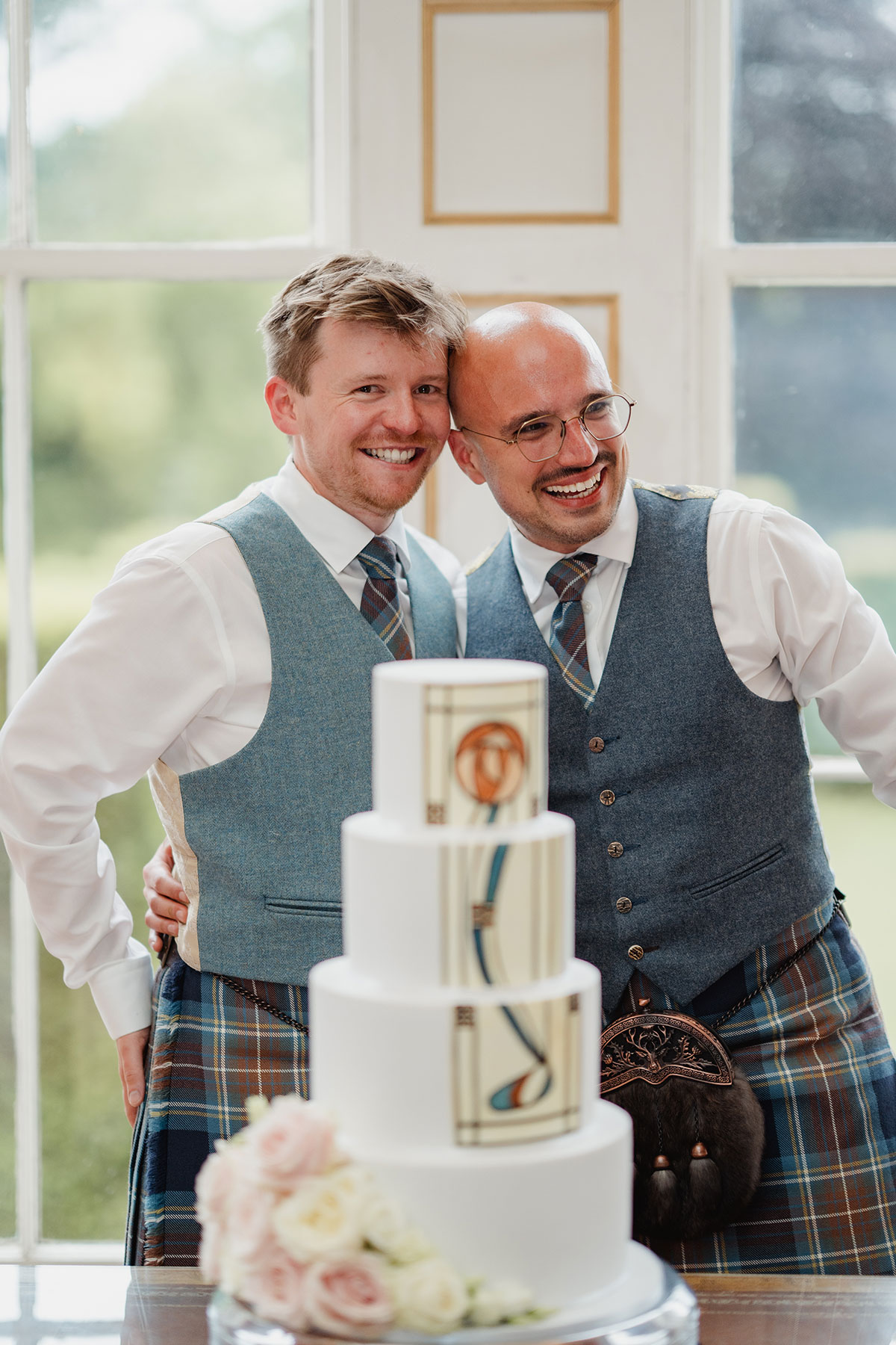 Two grooms dressed in matching waistcoats and tartan kilts smile and embrace beside a four-tier wedding cake decorated with geometric designs and topped with fresh roses