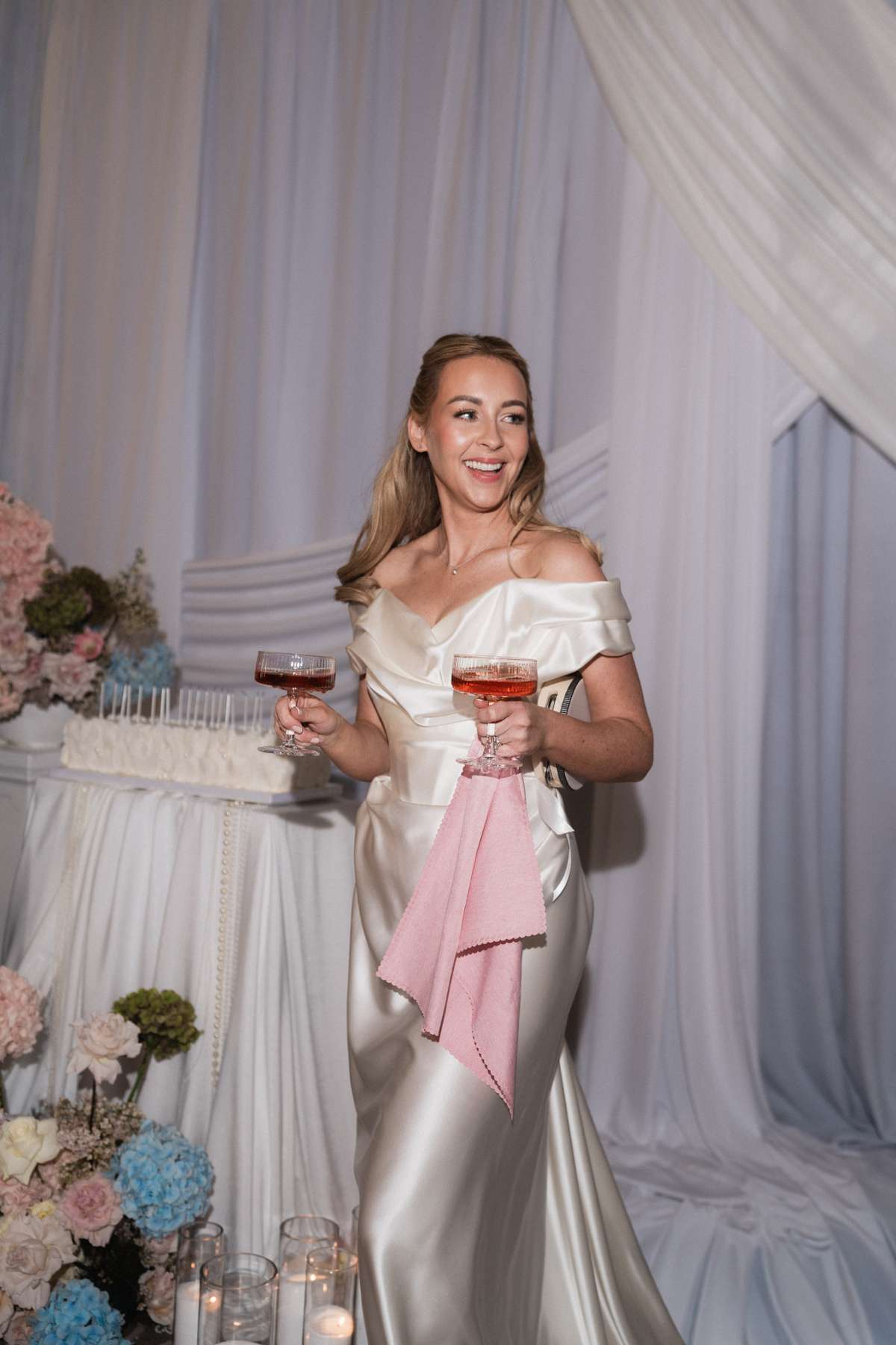 Bride Siân holds two glasses of pink champagne beside her long rectangular wedding cake at the Mar Hall reception.