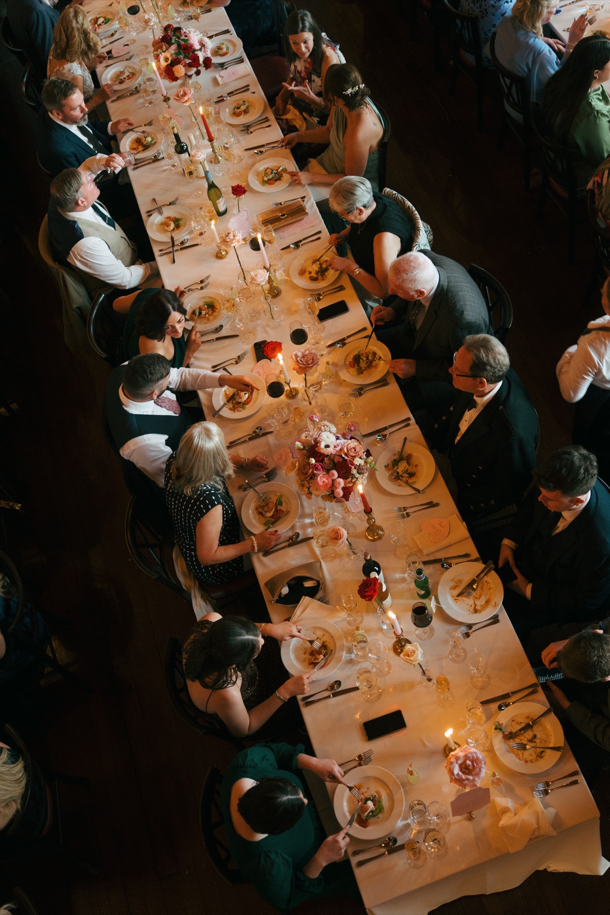 Aerial view of wedding guests enjoying dinner at Barnbougle Castle, with long tables decorated with pink-and-red flowers and candles.