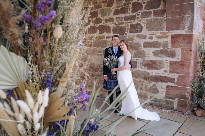 Bride and groom pose against a brick wall with dried flowers in shot