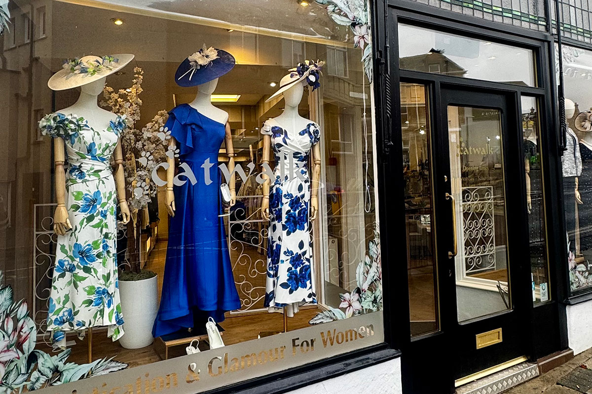 Catwalk boutique storefront with window display featuring mannequins in blue and floral occasion dresses and coordinating hats