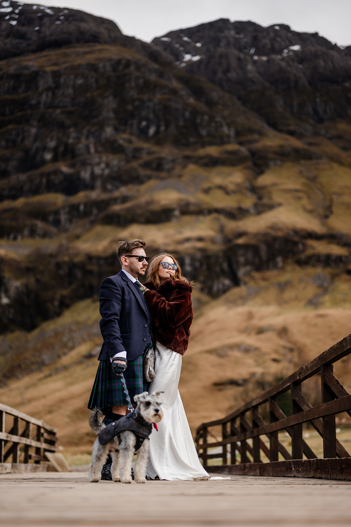 Bride and groom with their schnauzer dog on a wooden bridge in Glencoe, wearing sunglasses against the dramatic Highland backdrop