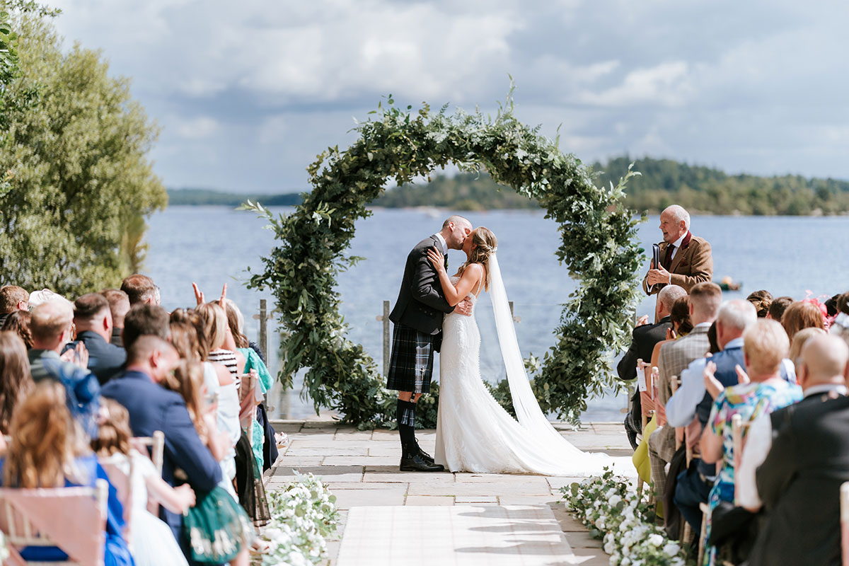Outdoor wedding ceremony at Lodge on Loch Lomond with couple kissing beneath floral arch and loch backdrop