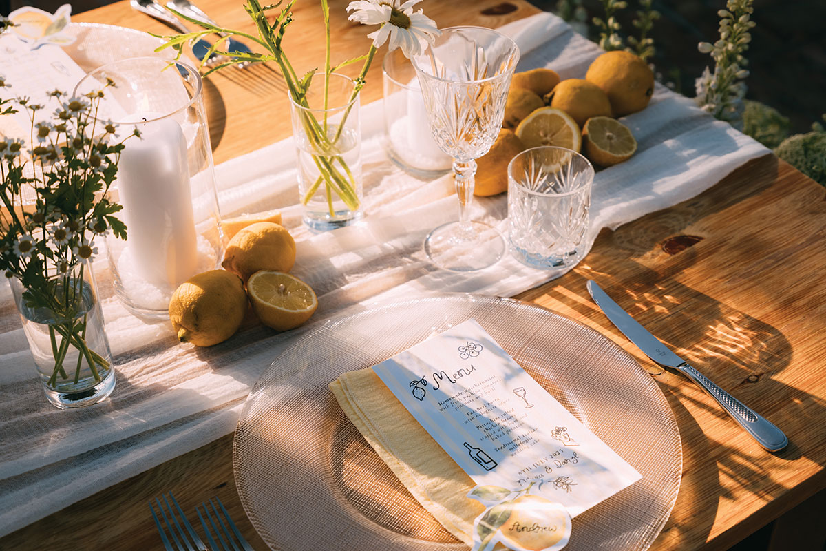 Close-up of a wooden wedding table styled with lemons, glassware, candles and a printed menu placed on a textured charger plate in warm sunlight