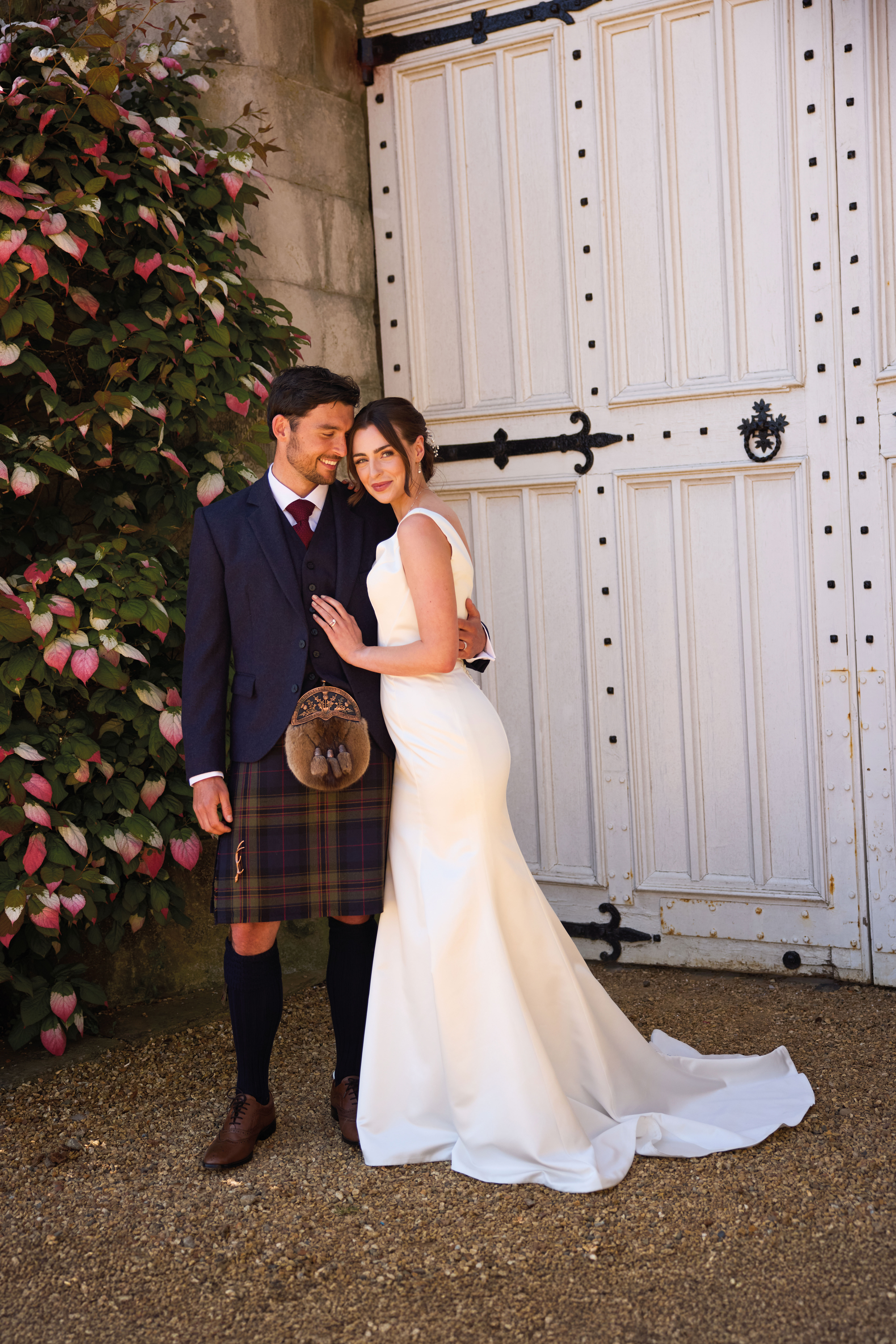 groom smiles at bride in front of white door at dundas castle for McCalls Highlandwear Pride of Noble tartan photoshoot