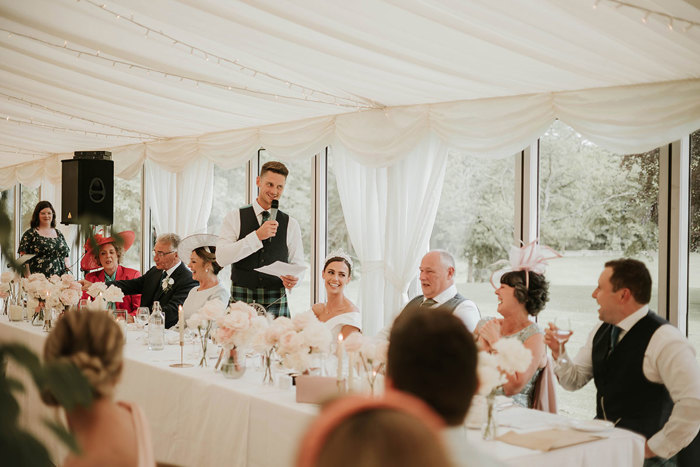 A groom standing up at the top table giving a speech holding a microphone and piece of paper while the guests laugh 