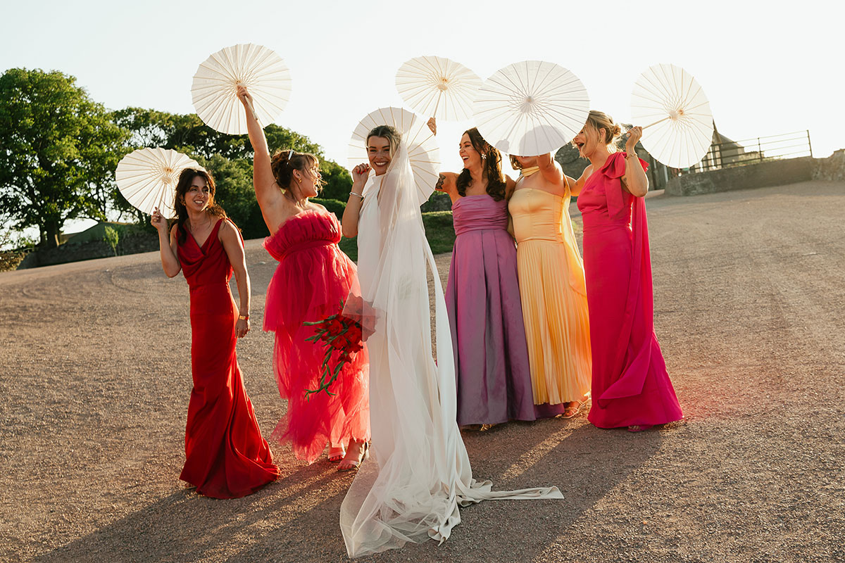 Bride and bridesmaids pose with white parasols during golden hour at Kinkell Byre, showcasing colourful pink, red, yellow and lilac dresses.