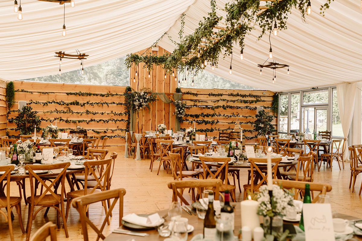 Rustic marquee reception with wooden tables and chairs, hanging greenery, and string lights under a white draped ceiling