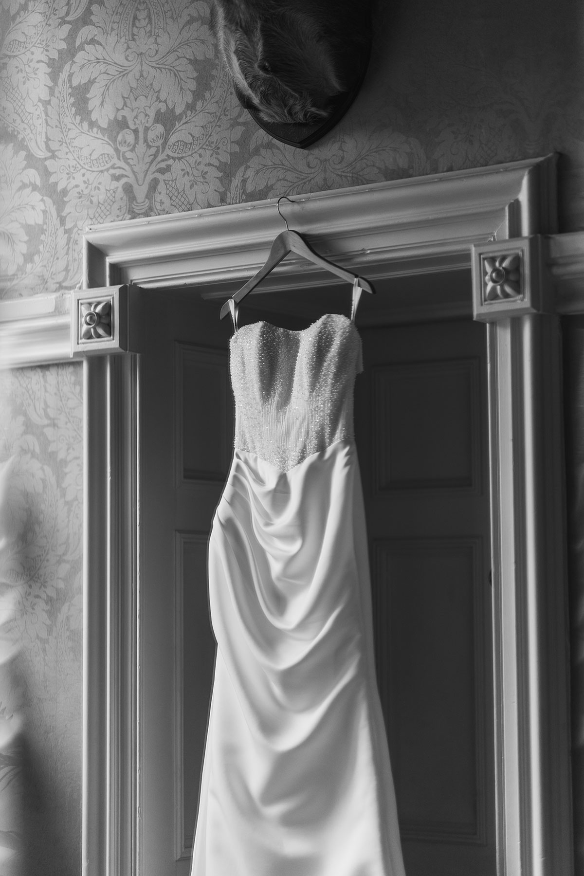 Black and white detail photograph of the bride's beaded strapless wedding dress hanging in the doorway during preparations