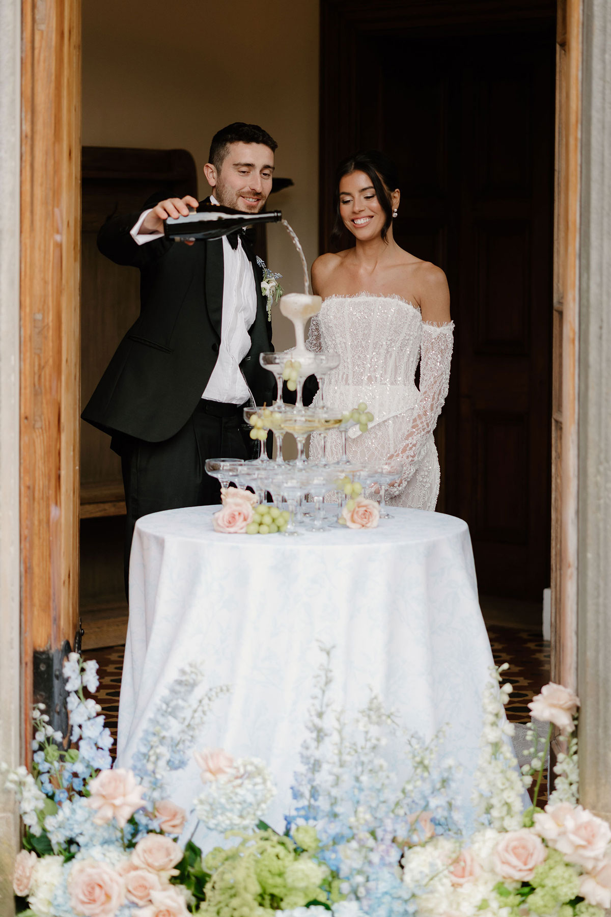 Bride and groom pouring champagne into a glass tower framed by pastel floral arrangements