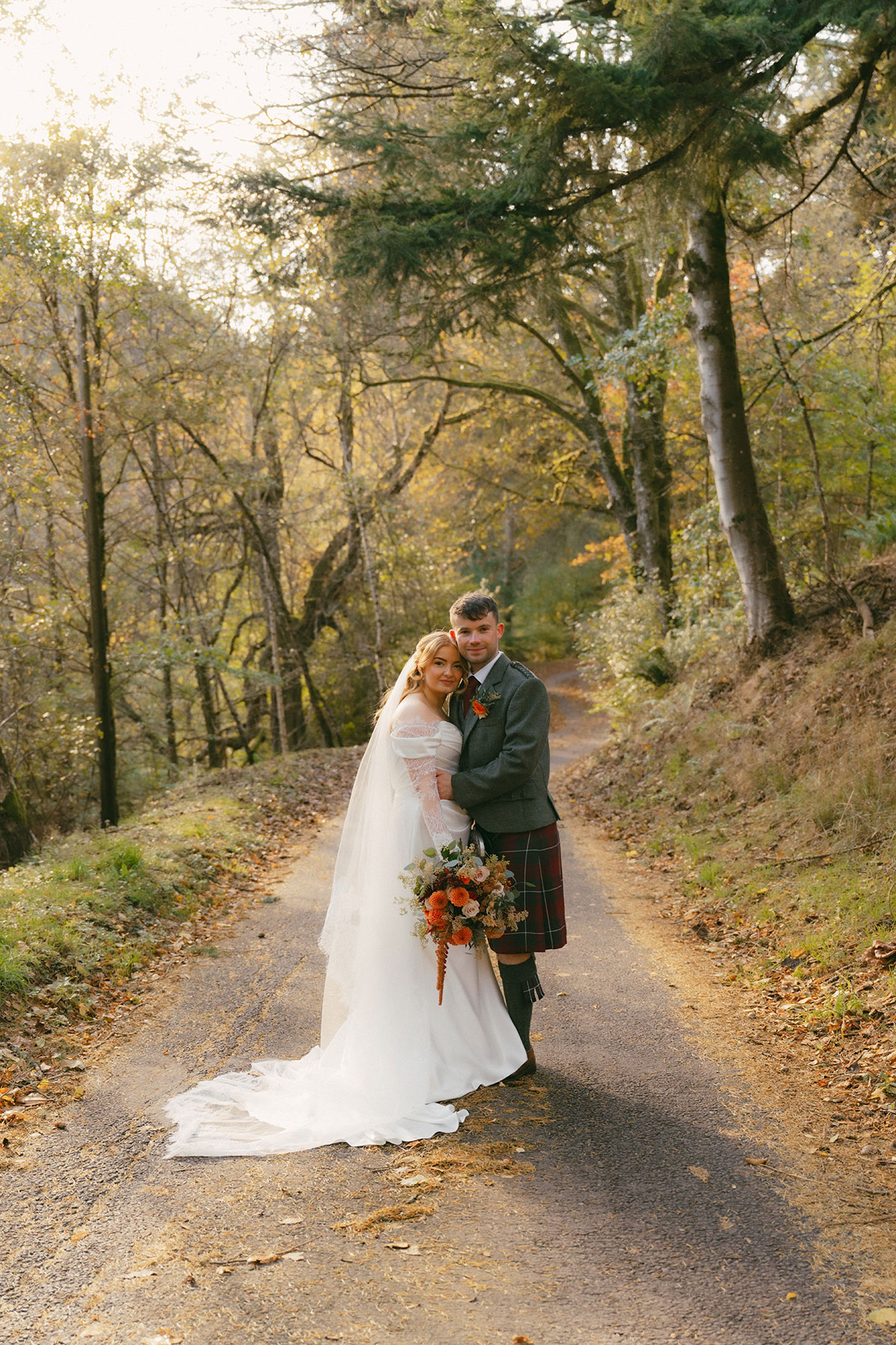 Bride and groom posing on tree-lined forest path with autumn leaves and bouquet at Scottish wedding