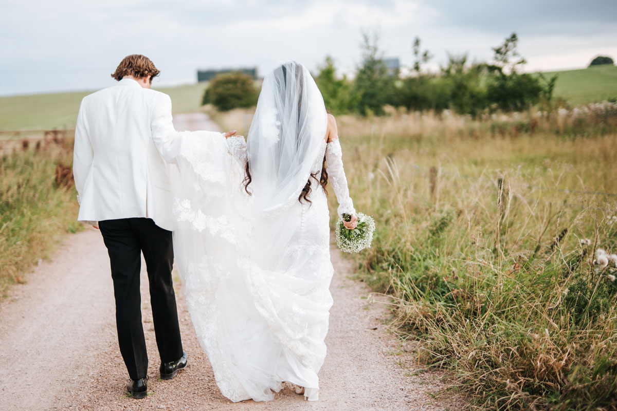Bride and groom walking away down a country track, bride holding bouquet and veil flowing behind