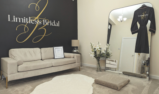 dressing room with beige carpet and couch, with flowers reflected in mirror that has black robe hanging on it. 'Limitless Bridal' is on the wall in gold and white writing. 