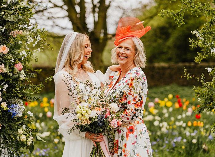 a smiling bride looks at a person wearing and orange and ivory floral patterned dress. They are standing in a garden carpeted with colourful flowers, tulips and daffodils