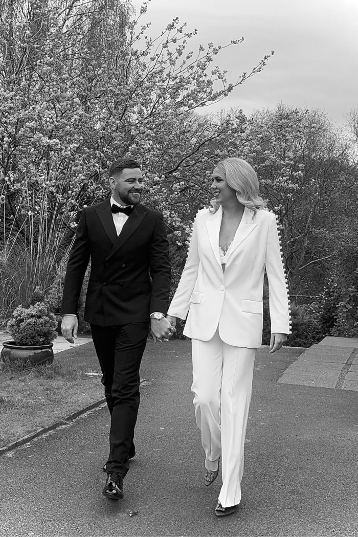 a bride and groom both wearing suits walk hand in hand through a garden in this black and white photo