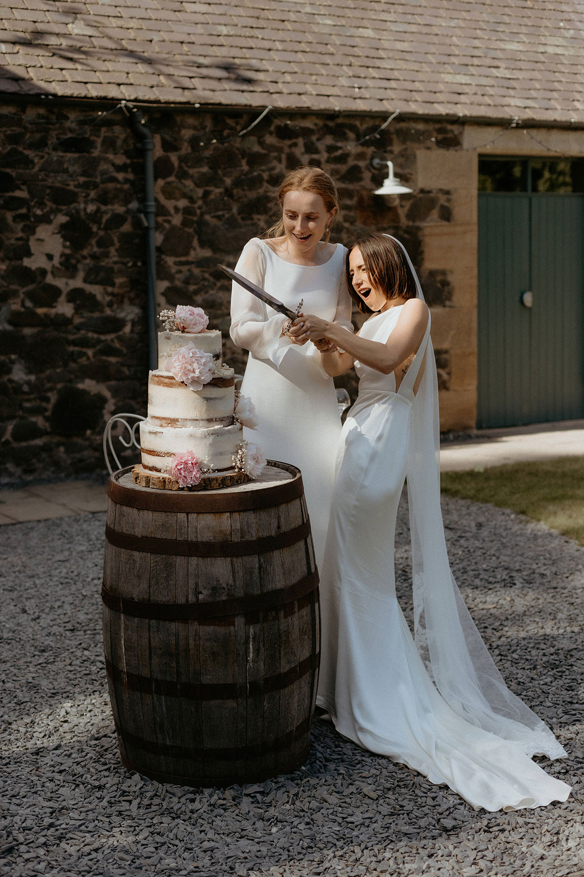 two brides cutting a semi naked buttercream wedding cake sitting on a wooden barrell with a sword in a stone courtyard setting