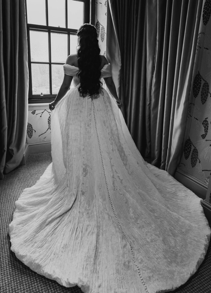 A bride facing away from the camera looking out of a window with her wedding dress train flowing over the floor behind her